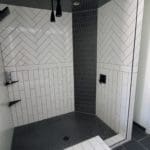 Bathroom remodeling project by CNT Builders in Spearfish, SD featuring a modern walk-in shower with white subway tile in vertical and herringbone patterns, contrasted by dark hex tile on the ceiling and floor, with matte black fixtures and built-in shelving.