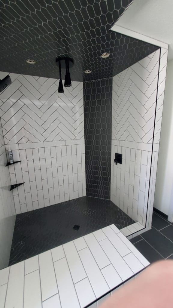 Bathroom remodeling project by CNT Builders in Spearfish, SD featuring a modern walk-in shower with white subway tile in vertical and herringbone patterns, contrasted by dark hex tile on the ceiling and floor, with matte black fixtures and built-in shelving.