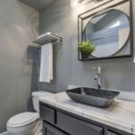 Modern bathroom remodel featuring a sleek black vanity with marble countertop, a vessel sink, matte black faucet, round mirror with geometric frame, and wall-mounted lighting. The space includes gray walls, a white toilet, and a metal towel rack with white towels.