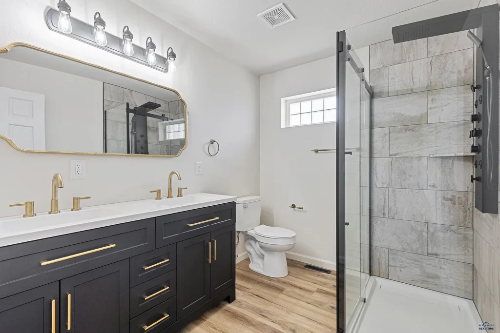 Modern bathroom remodeled by CNT Builders in Spearfish, SD, featuring a black double vanity with gold hardware and faucets, large framed mirror, walk-in shower with tile surround and glass door, wood-look flooring, and white toilet beneath a frosted glass window.
