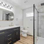 Modern bathroom remodeled by CNT Builders in Spearfish, SD, featuring a black double vanity with gold hardware and faucets, large framed mirror, walk-in shower with tile surround and glass door, wood-look flooring, and white toilet beneath a frosted glass window.