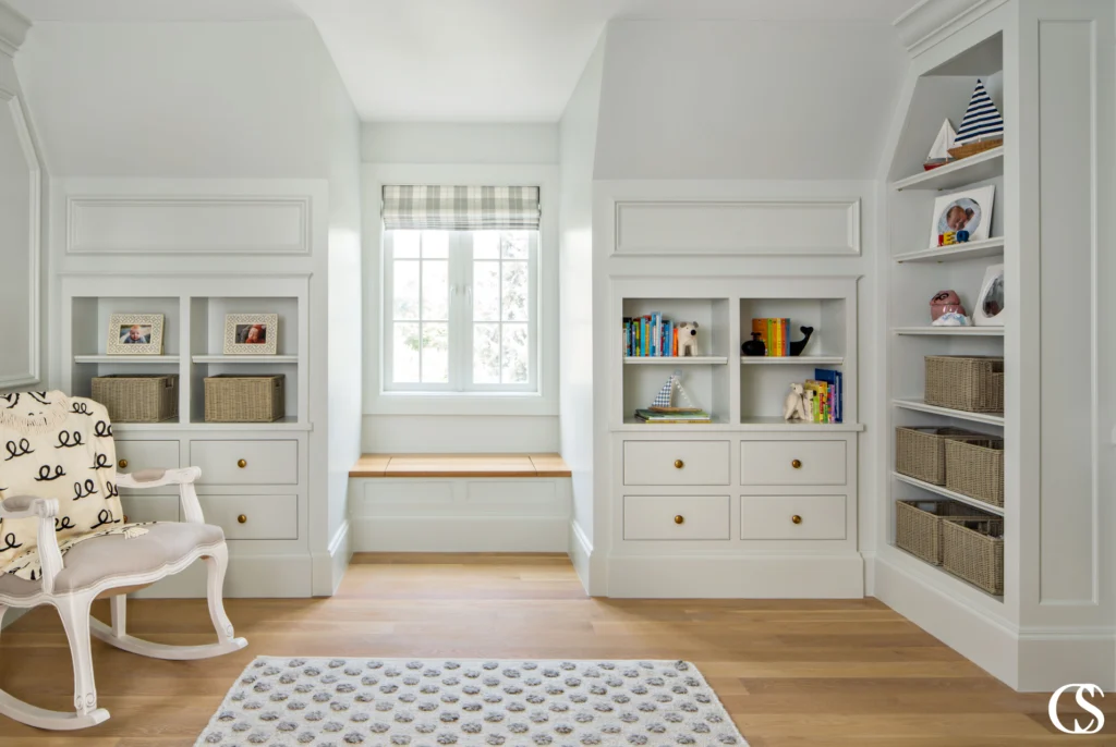 Custom built-in shelves with drawers, wicker baskets, and children's books flank a window bench with plaid Roman shade, designed by CNT Builders in Spearfish, SD.