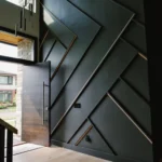 Modern entryway with a large pivot door and dramatic black geometric wall paneling, featuring angular patterns and natural light built by CNT Builders in Spearfish, SD.