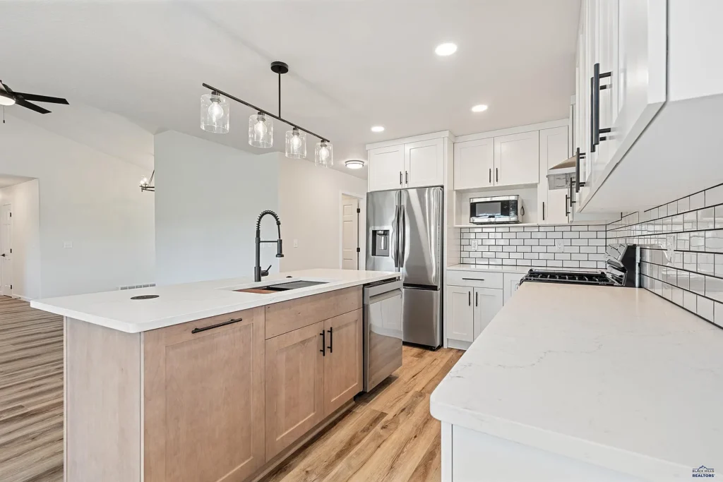 Modern kitchen remodeled by CNT Builders in Spearfish, SD, featuring white shaker cabinets, subway tile backsplash, stainless steel appliances, quartz countertops, light wood island with black fixtures, and pendant lighting over a deep sink.