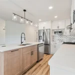 Modern kitchen remodeled by CNT Builders in Spearfish, SD, featuring white shaker cabinets, subway tile backsplash, stainless steel appliances, quartz countertops, light wood island with black fixtures, and pendant lighting over a deep sink.