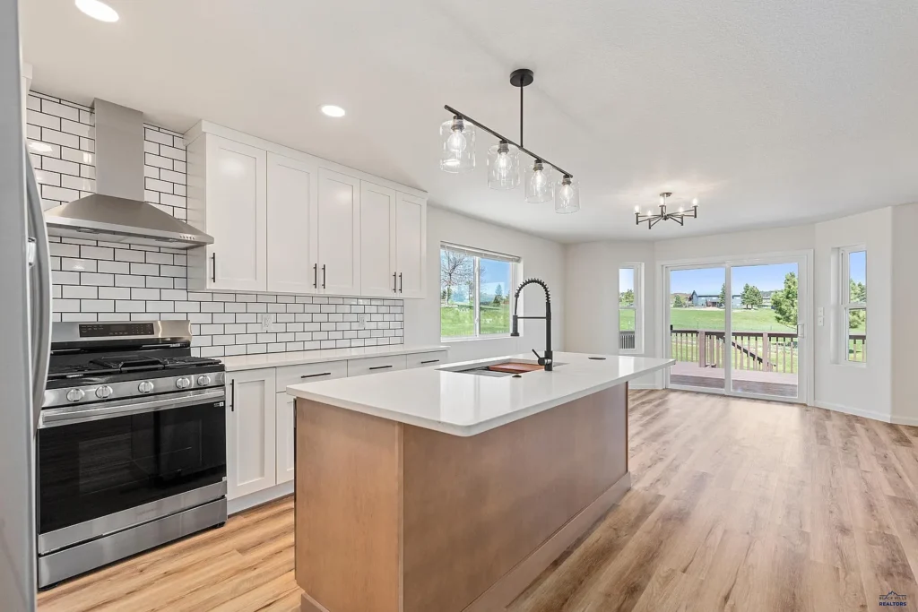 Bright, modern kitchen remodeled by CNT Builders in Spearfish, SD, featuring white shaker cabinets, subway tile backsplash, stainless steel gas range, quartz island with gooseneck faucet, pendant lighting, and sliding glass doors opening to a backyard deck with scenic views.