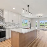 Bright, modern kitchen remodeled by CNT Builders in Spearfish, SD, featuring white shaker cabinets, subway tile backsplash, stainless steel gas range, quartz island with gooseneck faucet, pendant lighting, and sliding glass doors opening to a backyard deck with scenic views.