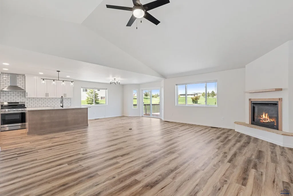 Open-concept living room and kitchen remodeled by CNT Builders in Spearfish, SD, featuring vaulted ceiling with ceiling fan, wood-look flooring, modern corner fireplace, large windows with scenic lawn views, white cabinets, subway tile backsplash, and stainless steel appliances.