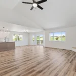 Open-concept living room and kitchen remodeled by CNT Builders in Spearfish, SD, featuring vaulted ceiling with ceiling fan, wood-look flooring, modern corner fireplace, large windows with scenic lawn views, white cabinets, subway tile backsplash, and stainless steel appliances.