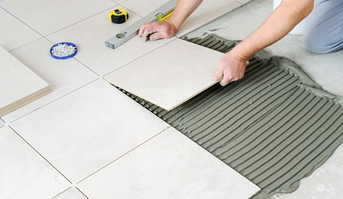 Tile installation in progress by CNT Builders in Spearfish, SD, showing a worker placing large beige ceramic tiles over thin-set mortar on a concrete floor.